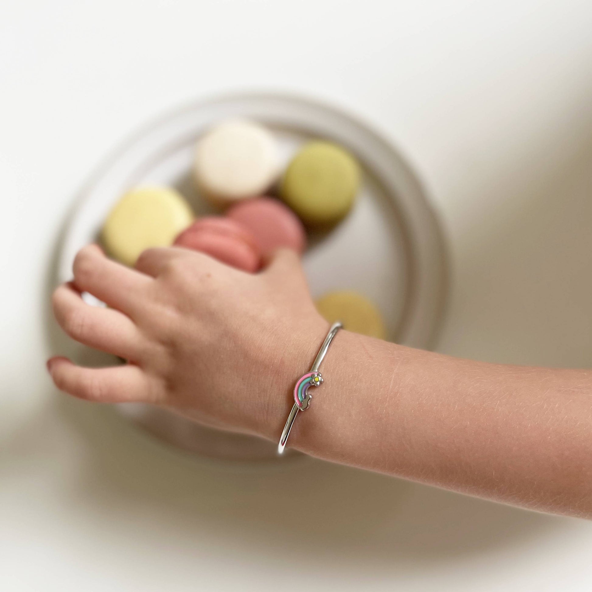 Little girl wearing Sterling Silver Pink Green and Purple Rainbow with Flower and Cloud on an Adjustable Bangle Cuff Bracelet reaching for macarons